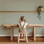 Smalle Strepen Lichtblauw – Atelier Chien: behang achter een kind aan een houten tafel.